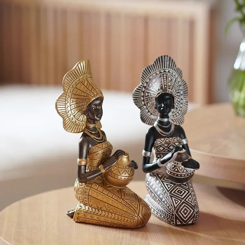 Two seated African resin woman statues with headpieces, one in gold and one in black-and-white tribal design, each holding a decorative pot
