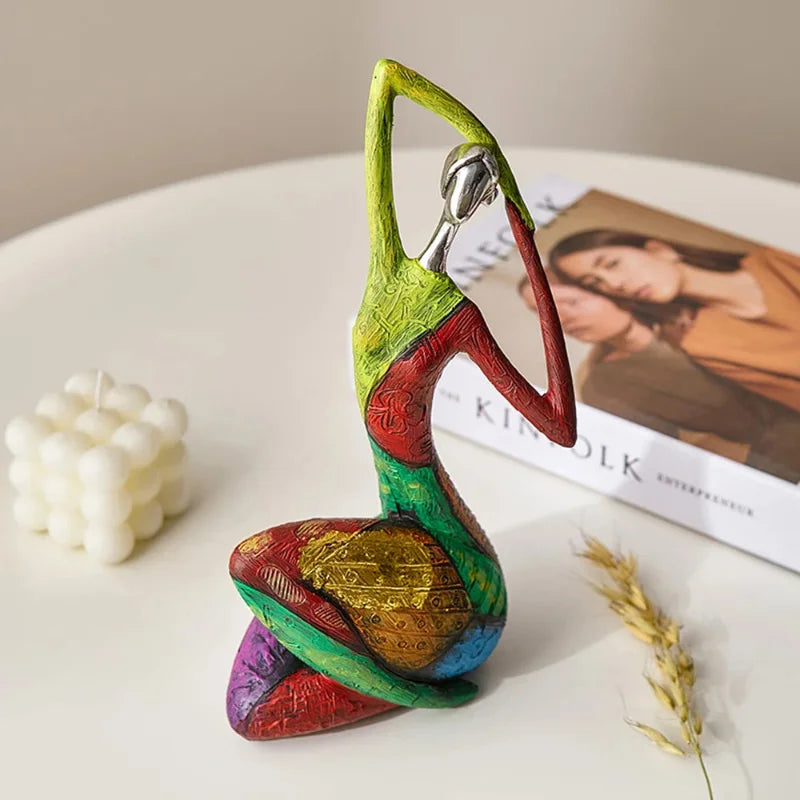 Kneeling colorful abstract resin statue with silver-look head and patchwork texture, posed with arms overhead on a white tabletop