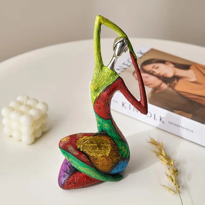 Kneeling colorful abstract resin statue with silver-look head and patchwork texture, posed with arms overhead on a white tabletop