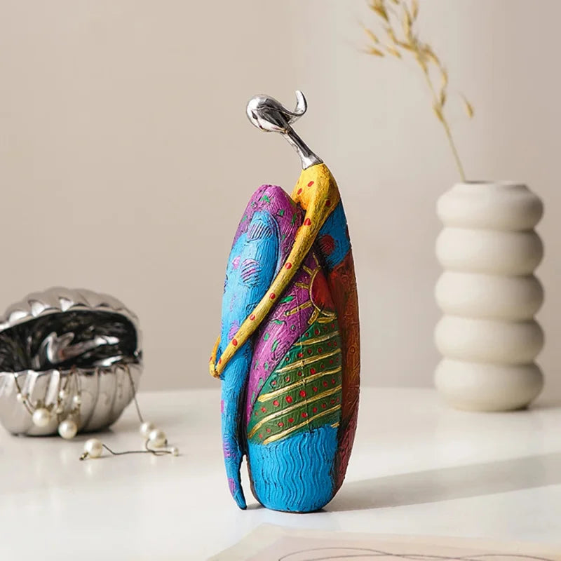 Colorful abstract resin statue in a wrapped pose with silver-look head, featuring blue, green, purple, and yellow patchwork patterns on a tabletop