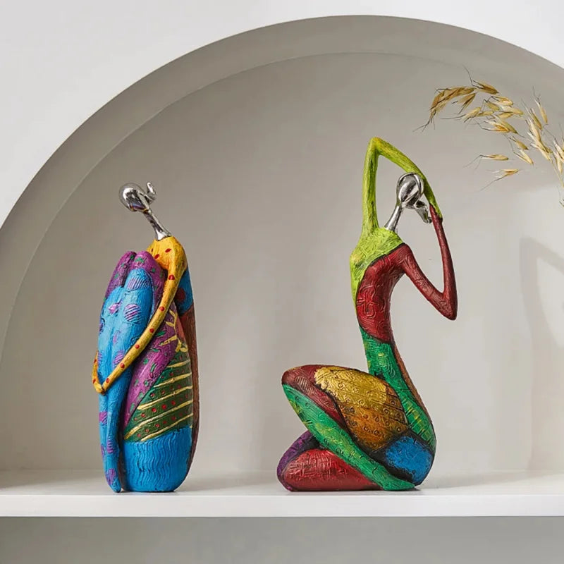 Pair of colorful abstract resin statues with silver-look heads, one standing in a wrapped pose and one kneeling with arms overhead, displayed on an arched white shelf