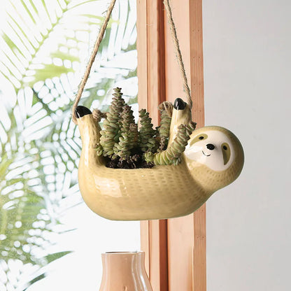 Yellow ceramic sloth hanging planter with trailing succulents, suspended by rope near a sunlit window with tropical foliage visible in the background.