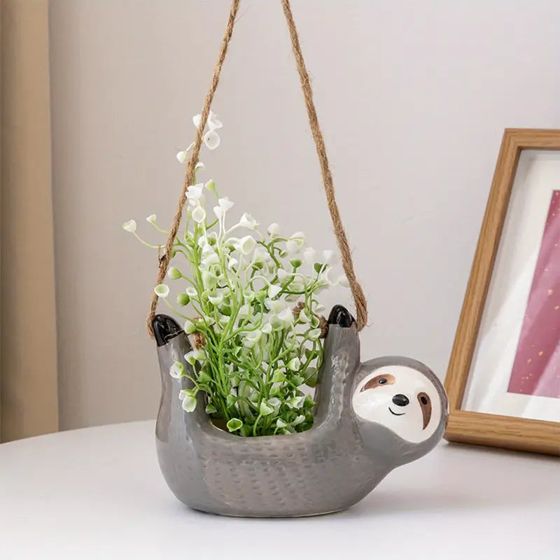Gray ceramic sloth hanging planter with white artificial flowers, displayed on a white table next to a framed picture in a cozy indoor setting.
