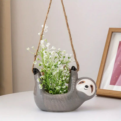 Gray ceramic sloth hanging planter with white artificial flowers, displayed on a white table next to a framed picture in a cozy indoor setting.